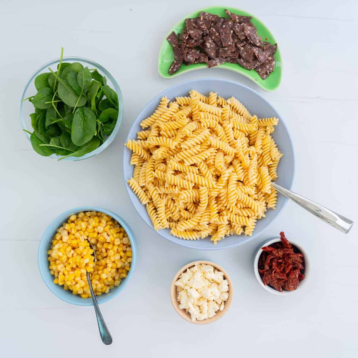 A bowl of dressed pasta surrounded by small bowls of feta, sundried tomatoes, corn, spinach leaves and salami.