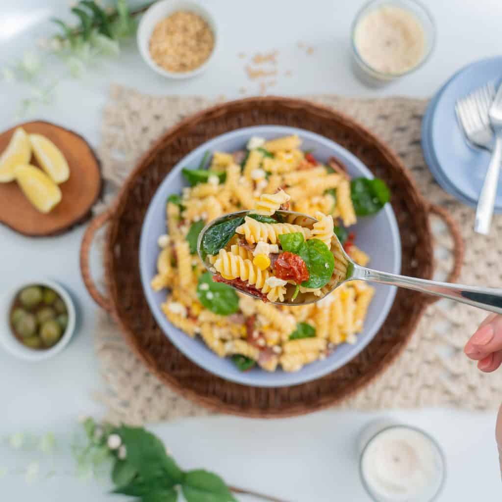 A serving spoon of Italian pasta salad being held above a large salad bowl, spiral pasta, sundried tomato, salami, corn kernels and feta crumbles visible.