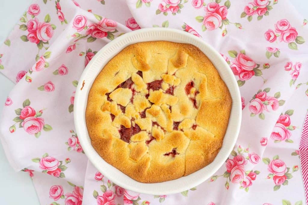 A golden brown pie with red filling in a pie dish sitting on a pink teat towel decorated with roses.