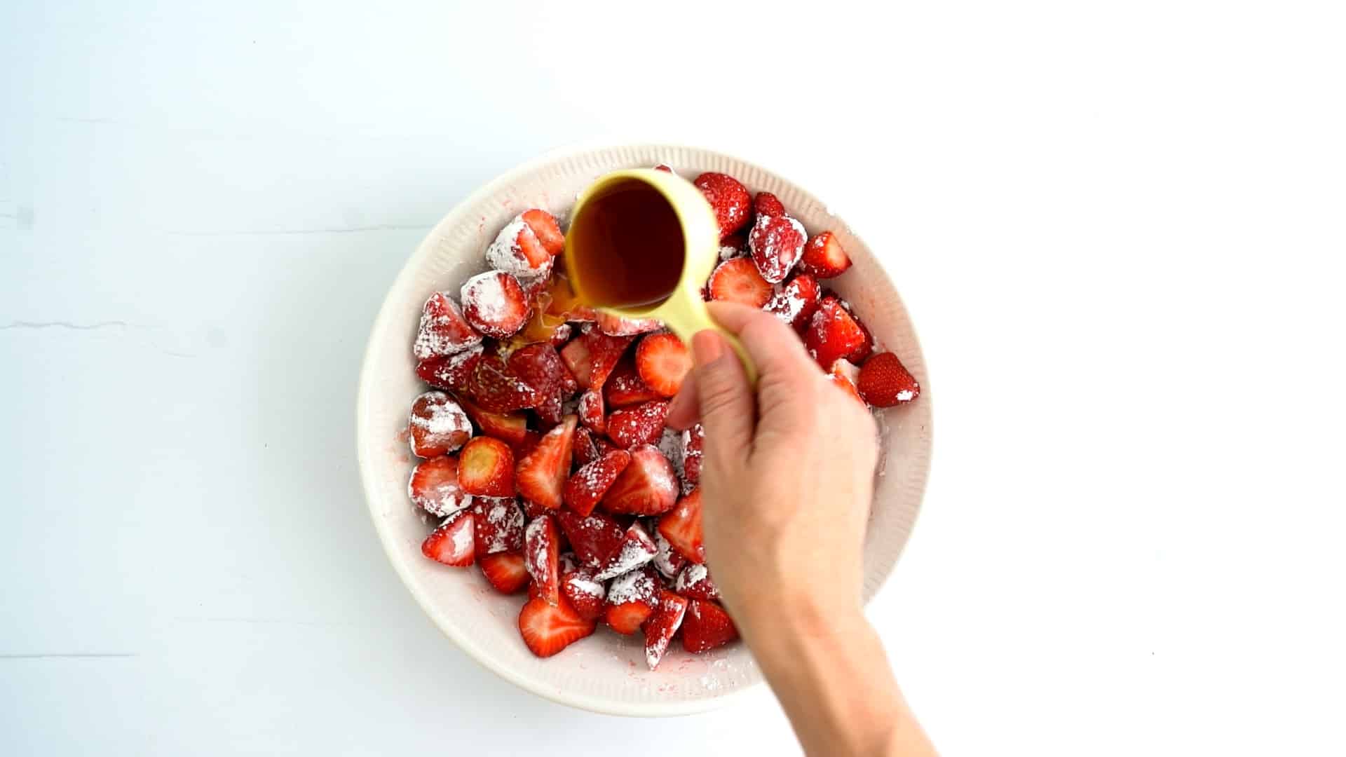 Strawberries dusted with cornflour in the bottom of a pie dish, a hand pouring maple syrup into the dish. 