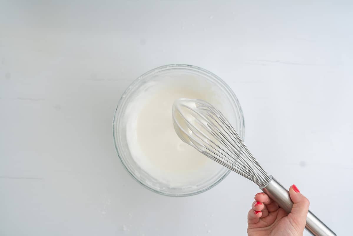 A whisk with smooth white icing dripping into a glass mixing bowl.