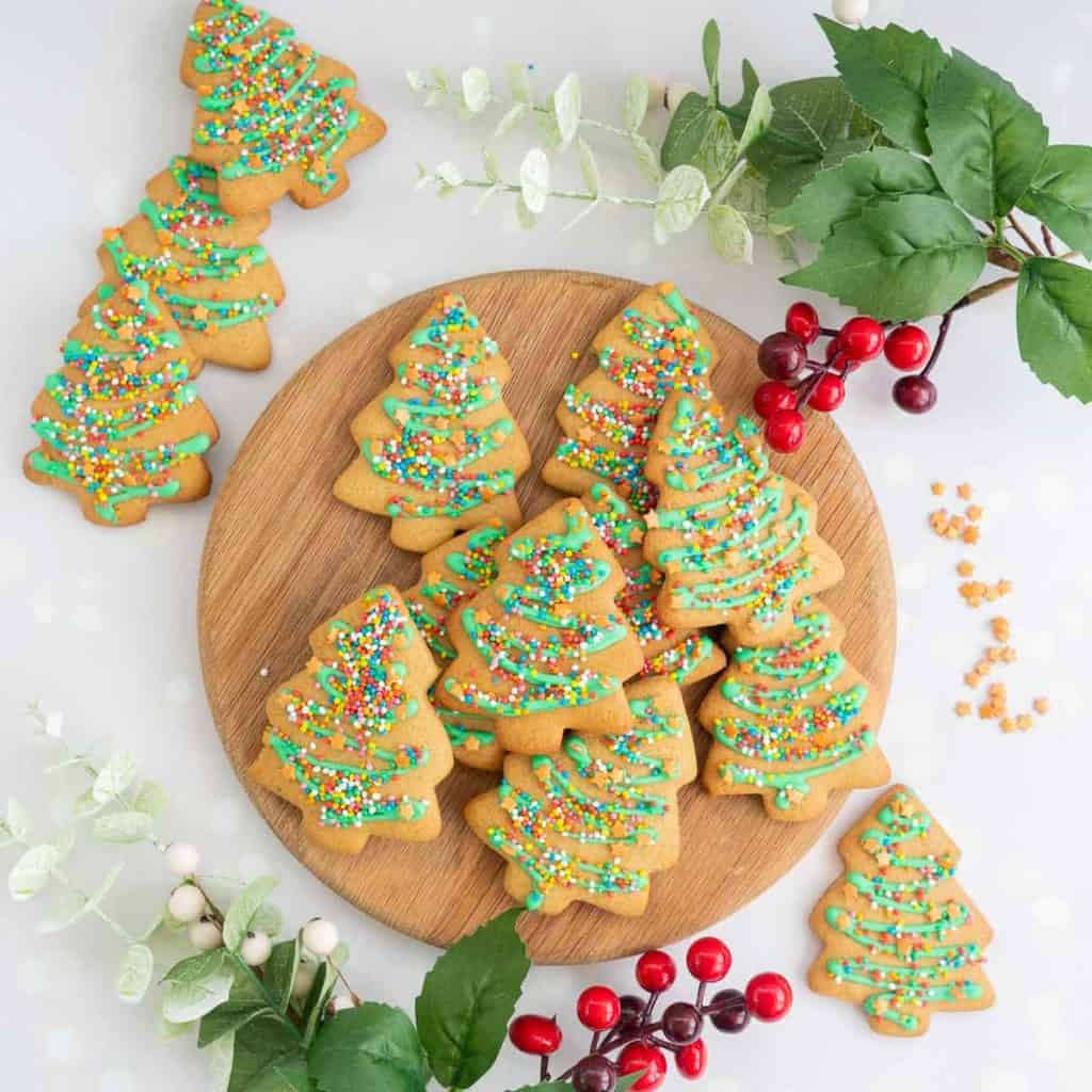 Christmas tree shaped cookies decorated with green icing and colourful sprinkles served on a round wooden serving board.