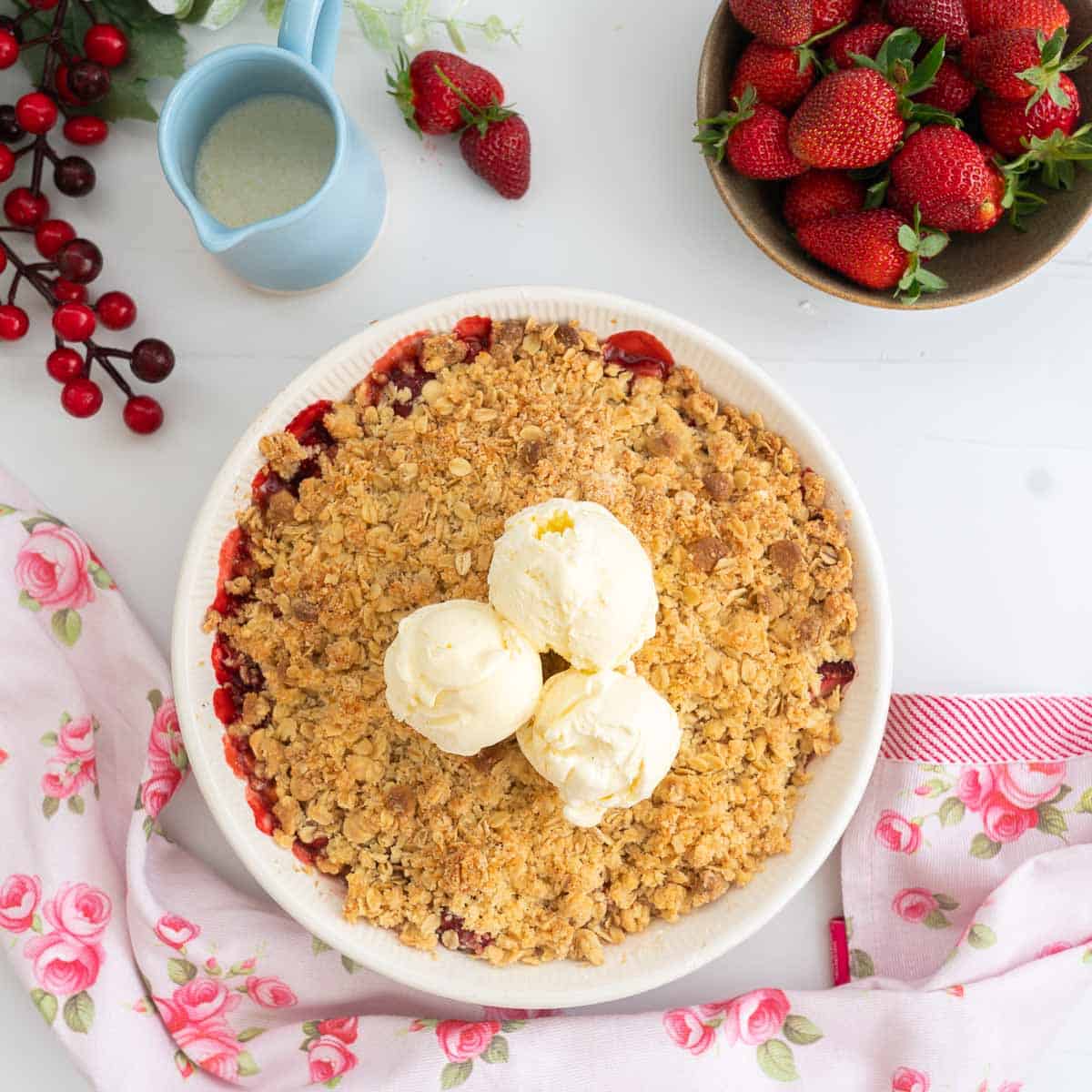 A fruit crumble topped with three scoops of ice cream on a white counter top with strawberries and a pink floral tea towel.
