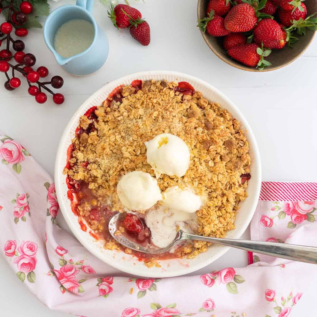 Image of a strawberry crumble, serving spoon and ice cream, strawberry syrup visible under the crumble topping.