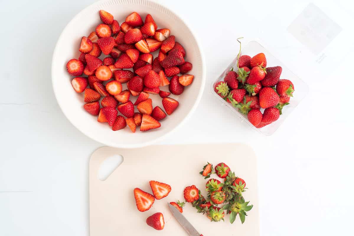 Strawberries and a knife on chopping board, a pie dish filled with hulled strawberries.