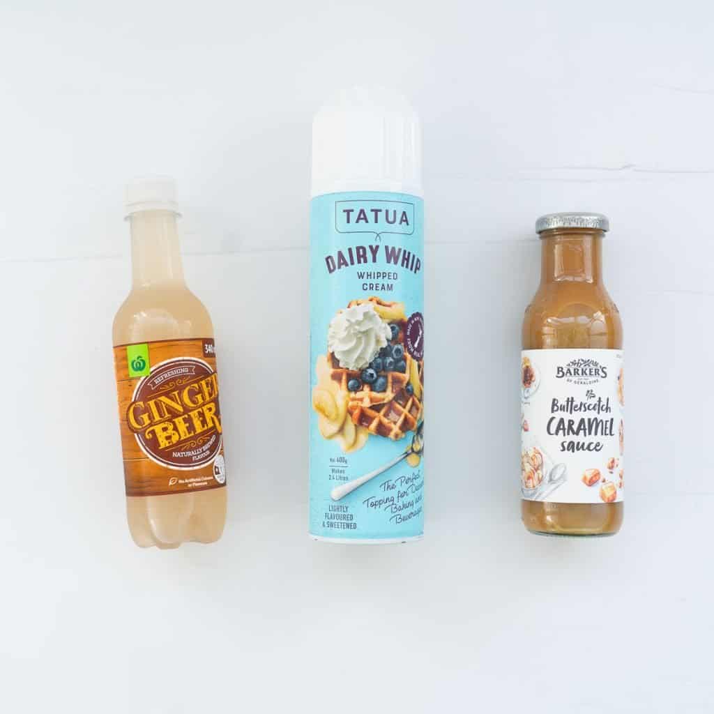 Ginger beer, butterscotch sauce and canned whipped cream lying on a white benchtop.