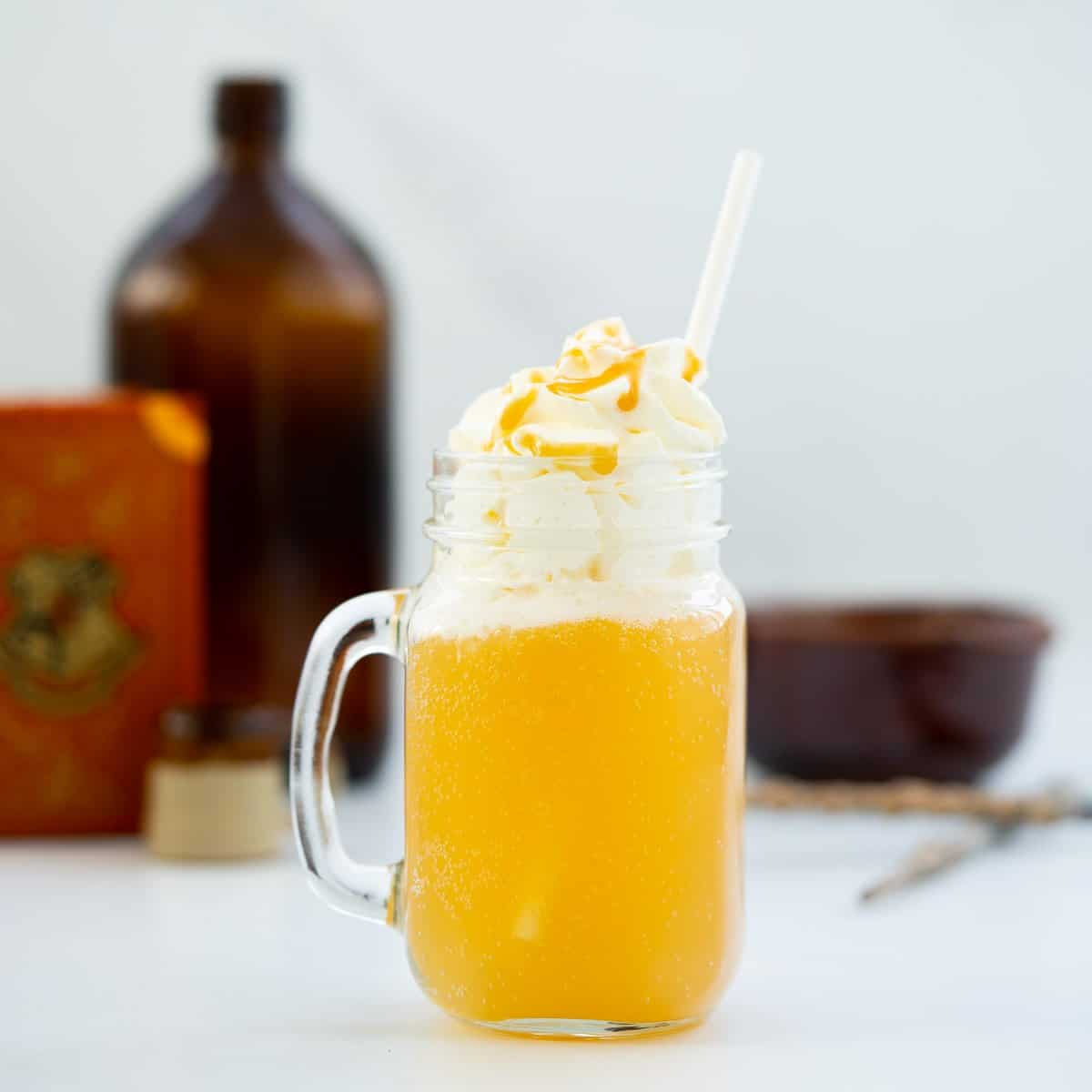 A glass beer handle filled with butterbeer and whipped cream, brown glass bottle and golden wands in the background. 