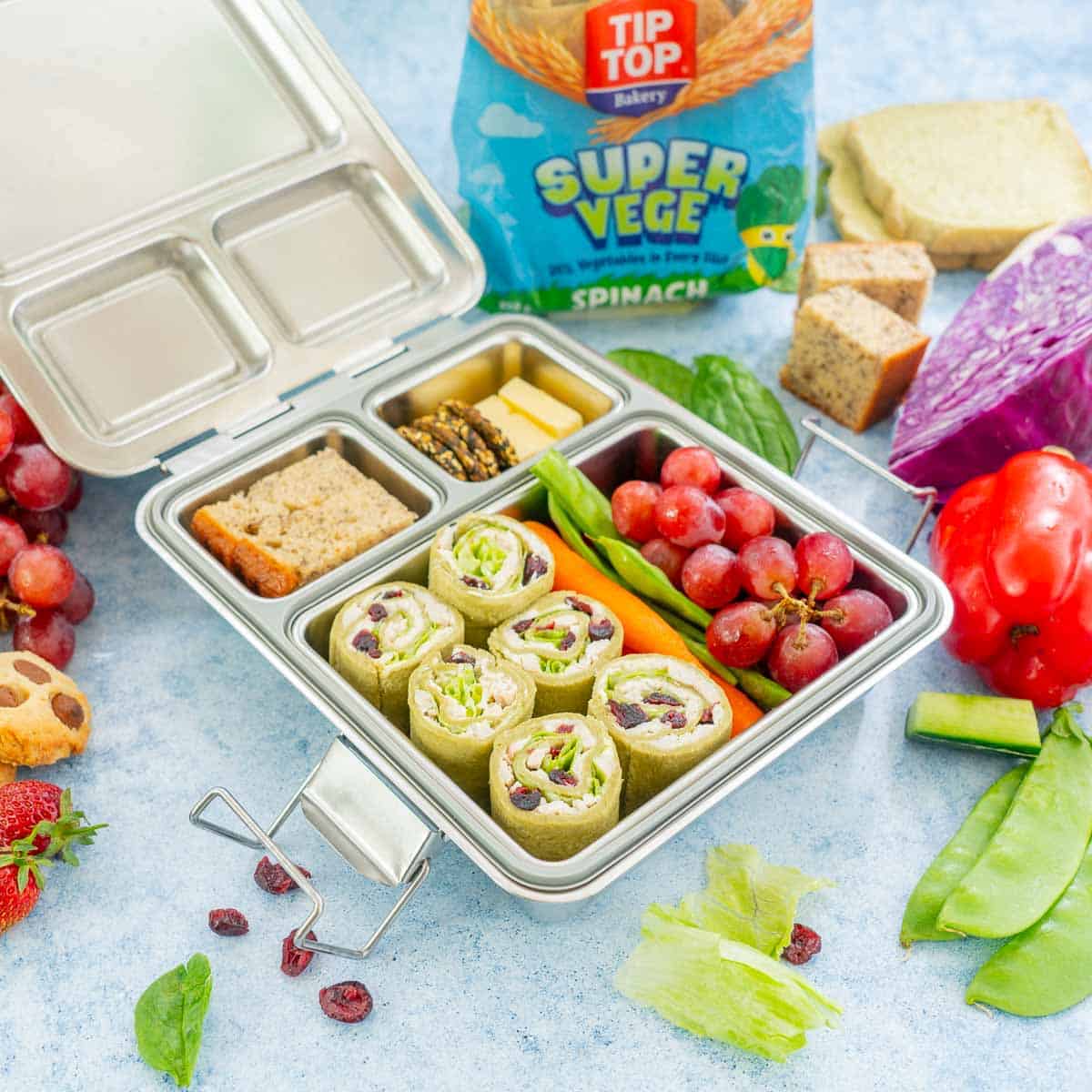 Stainless steel bento lunchbox packed with pinwheel sandwiches, fruit crackers, vegetables and banana cake.