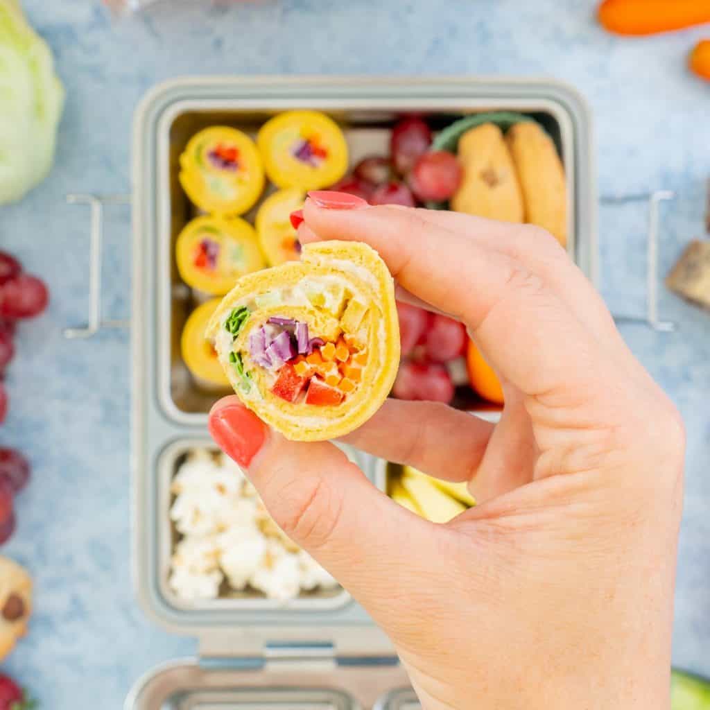 A rainbow vegetable pinwheel sandwich being held above a packed stainless steel lunchbox.