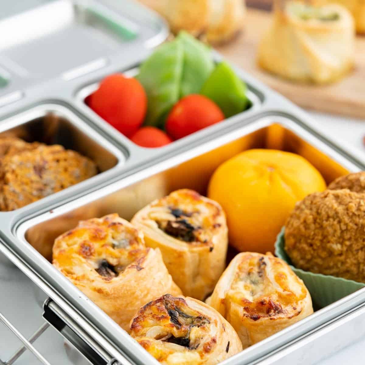 Chicken and alfredo puff pastry scrolls packed in a stainless steel bento lunchbox with colourful produce.