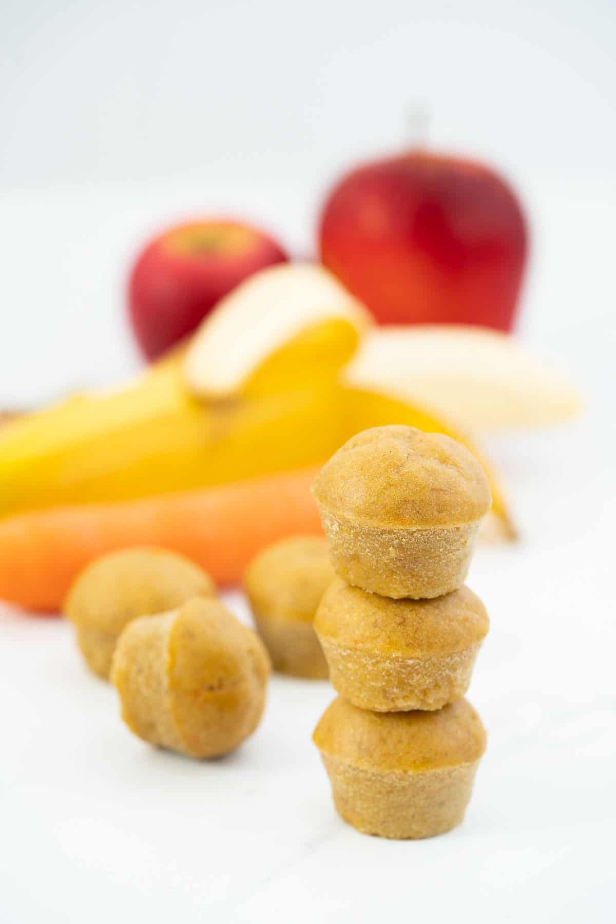 a tower of three mini muffins with apple, banana and carrots in the background.