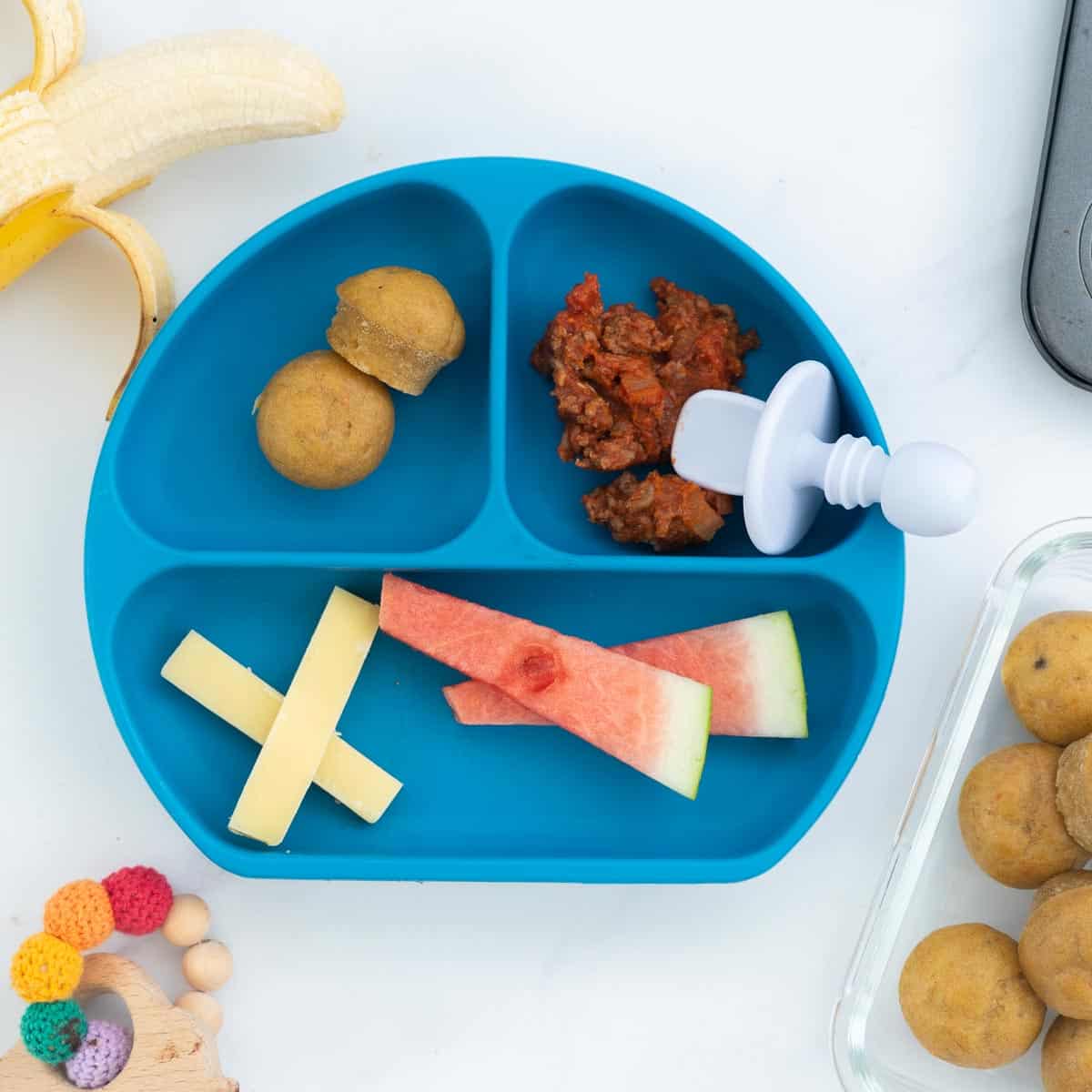 A blue silicone divided plate with a meal of cheese sticks, mini muffins, minced beef and watermelon.