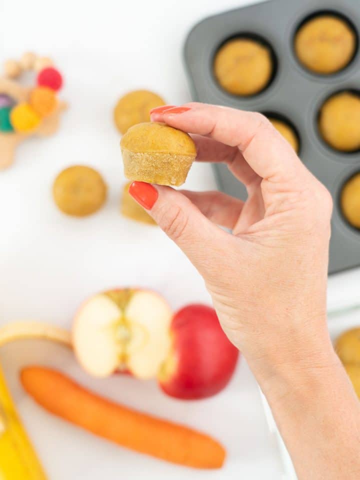 A women's hand holding a mini muffin above a tray of baked mini muffins.