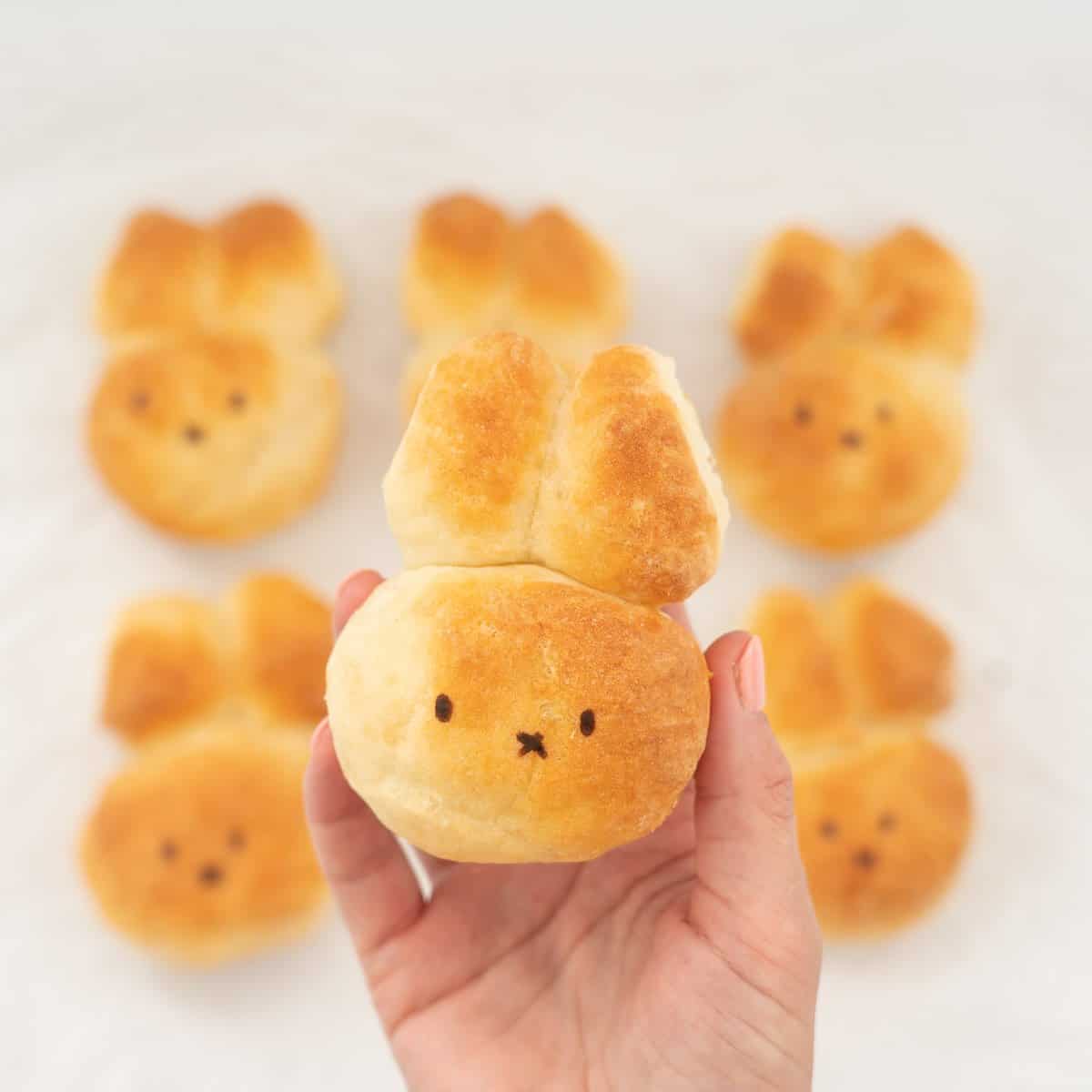 A bread roll decorated to look like a rabbit beng held above a tray of other golden baked rabbit breads.