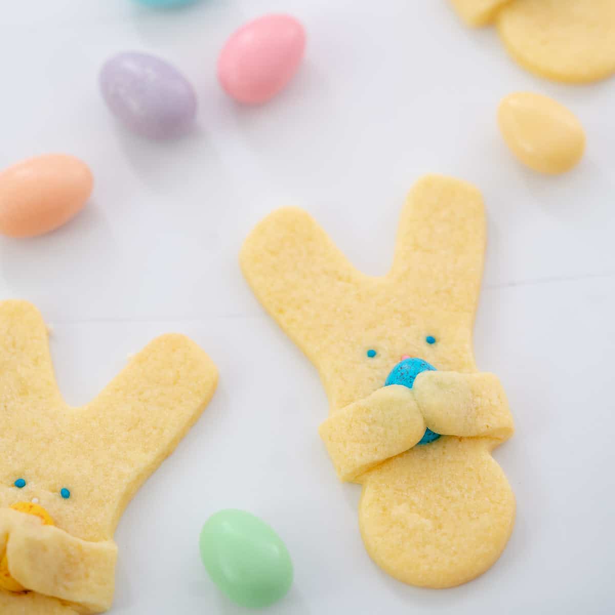 Easter bunny cookies holding mini chocolate easter eggs on a white bench top scattered with pastel coloured sugared almonds. 
