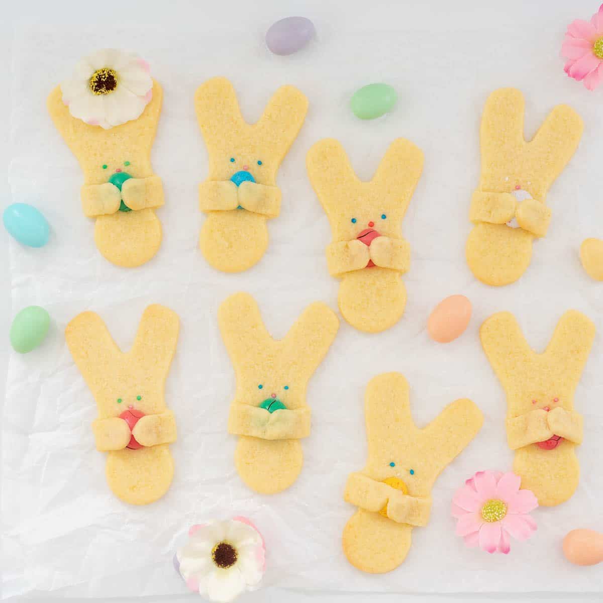 Bunny shaped sugar cookies holding a mini easter egg, on a white bench top scattered with pastel easter eggs and spring flowers.