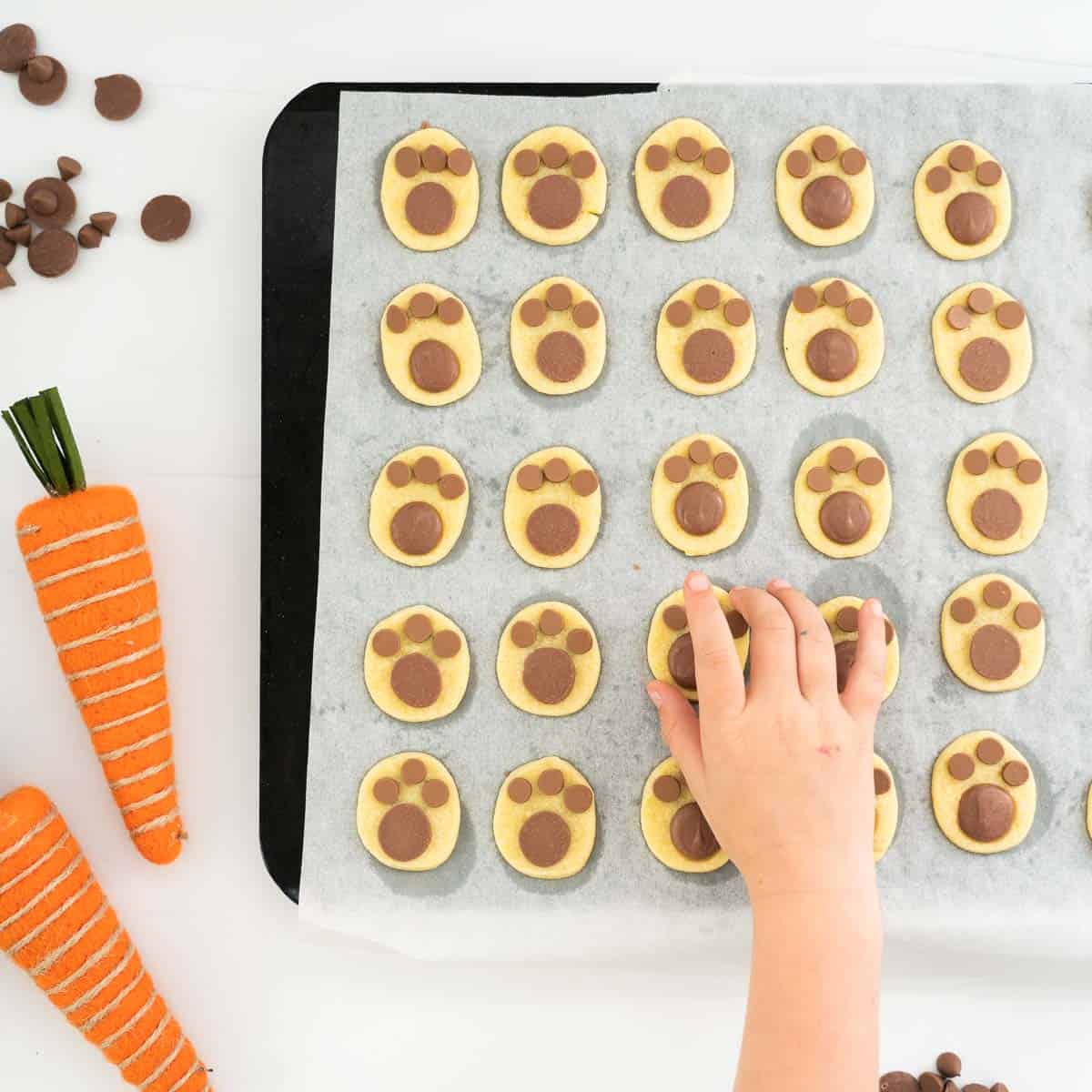 A child's hand reaching for a cookie off a baking sheet full of footprint shaped cookies.