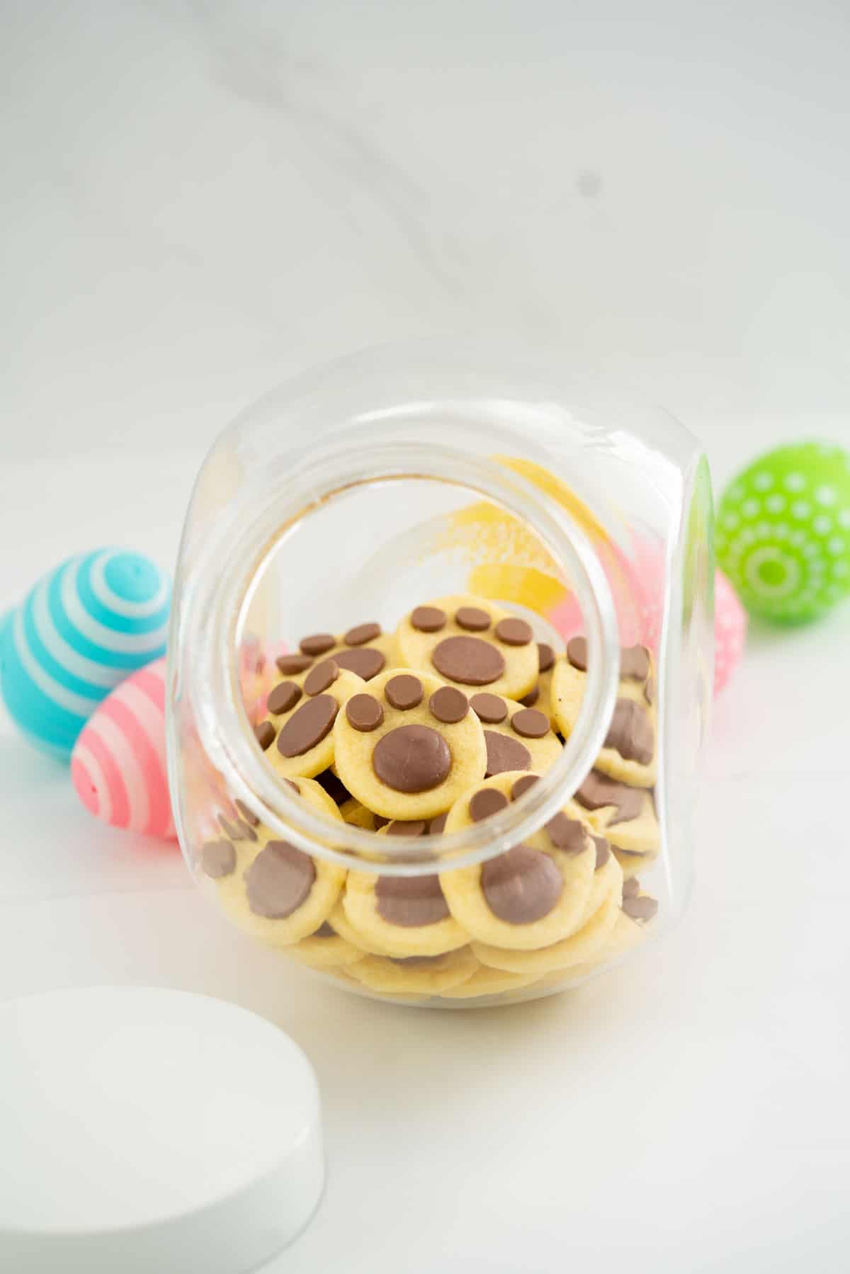 Bunny footprint cookies in a glass cookie jar, colourful easter eggs in the background.