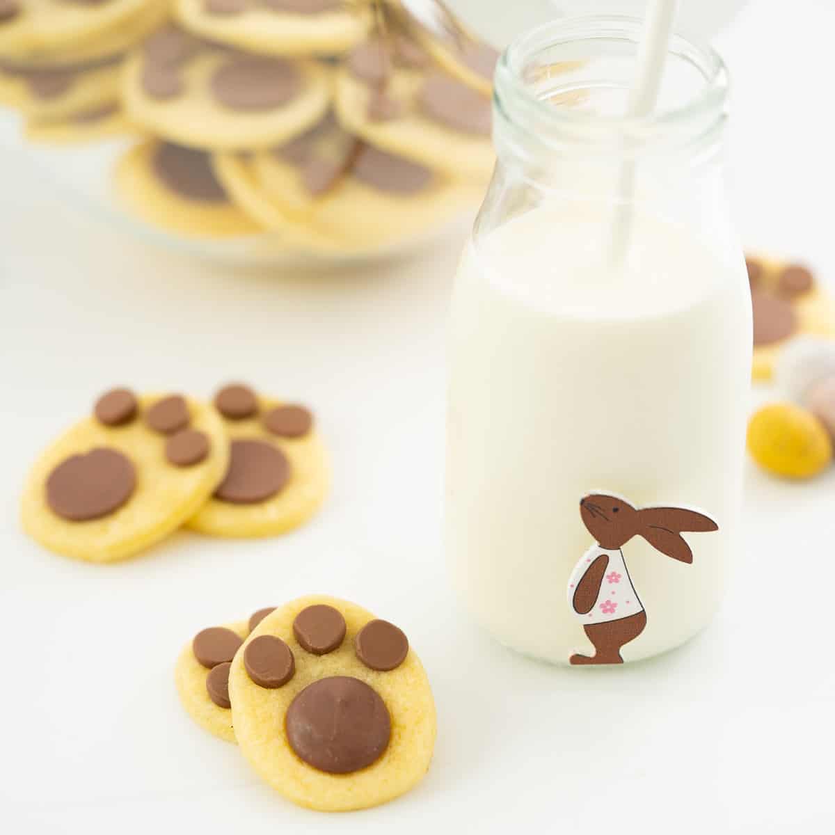 Bunny footprint cookies on a white bench top next to a small glass bottle of milk decorated with a picture of the easter bunny.