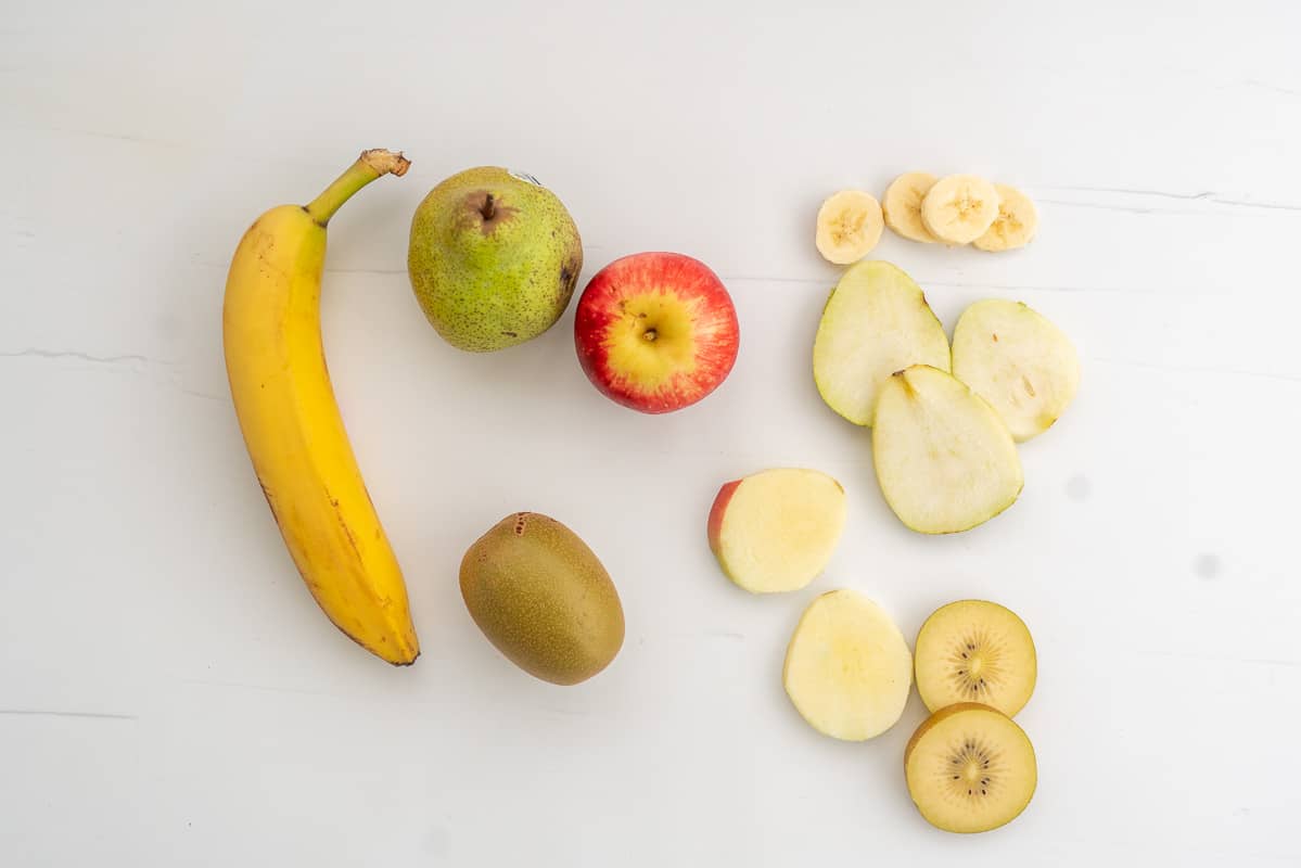 Banana, pear, apple and kiwifruit, whole fruit and slices of these fruit on a white bench top.