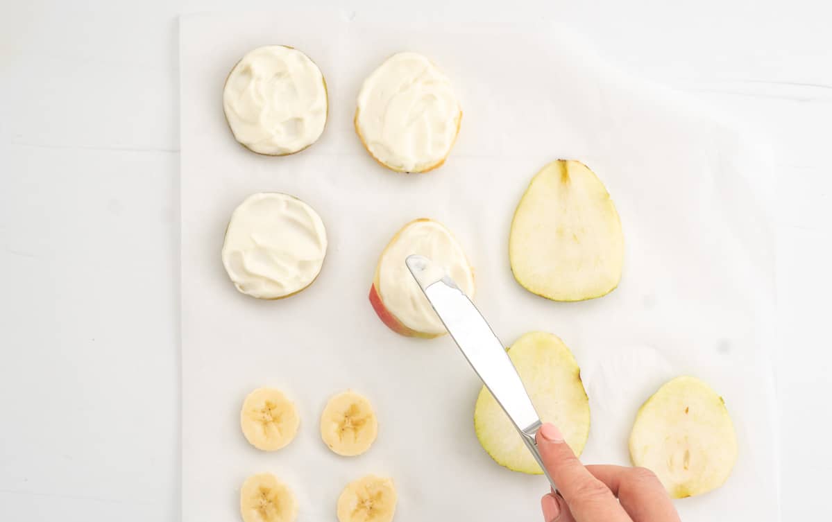 Slices of fruit on parchment paper being spread with thick yoghurt.