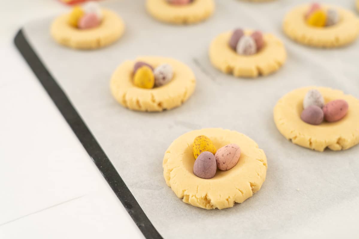 Vanilla nest cookies topped with 3 mini easter eggs ready to be baked on a baking sheet.