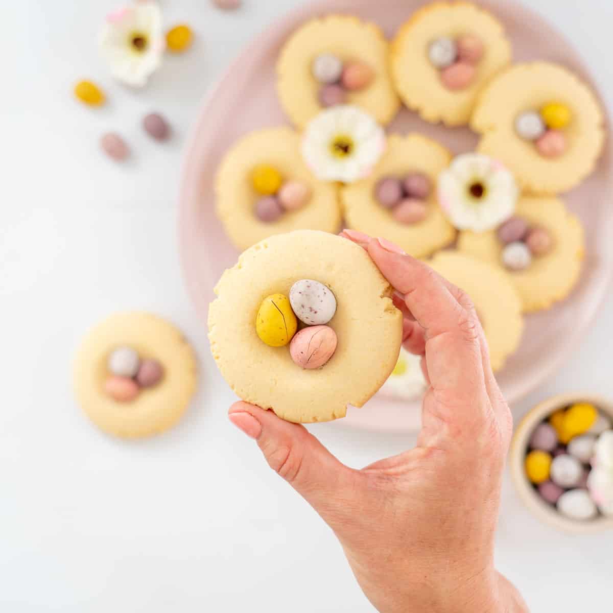 a vanilla cookie topped with 3 pastel mini eggs being held above a plate of cookies.