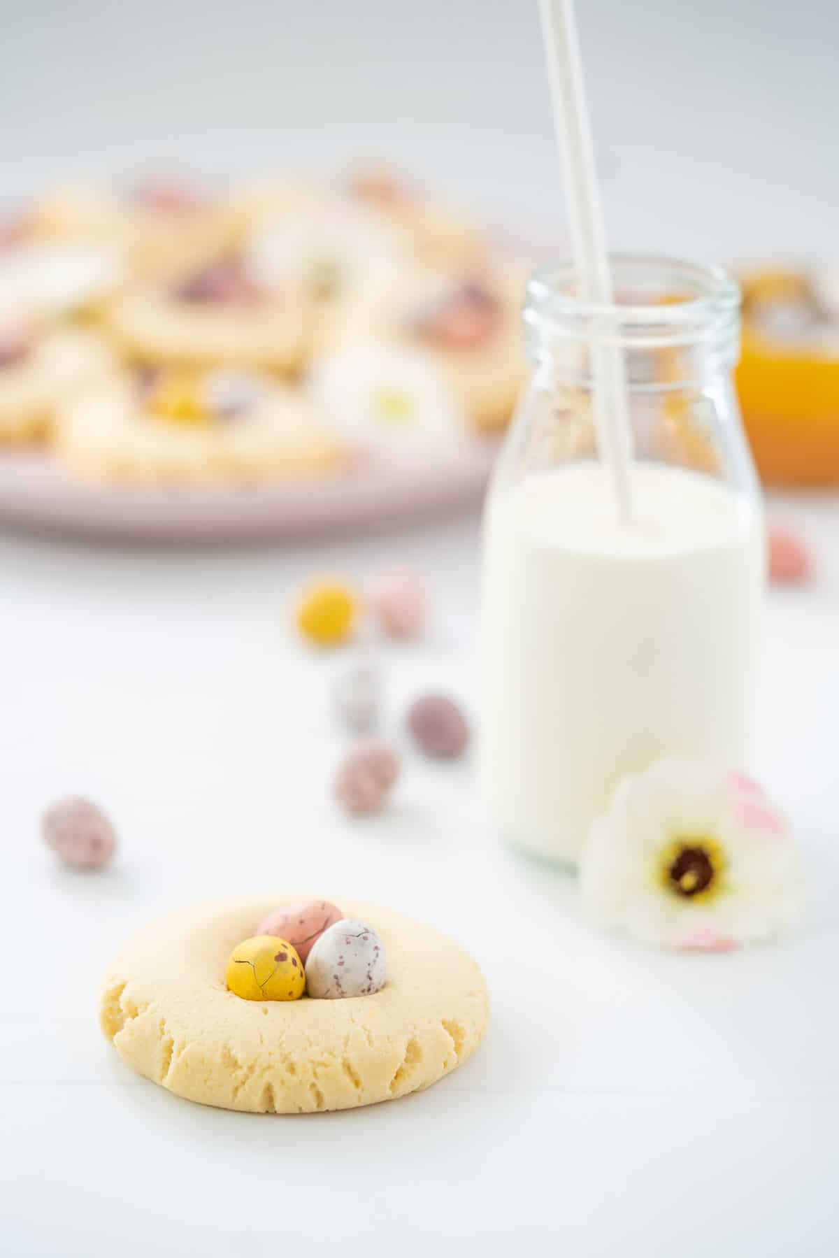 A mini egg cookie on a white bench top that is scattered with mini eggs and a small glass bottle of milk. 