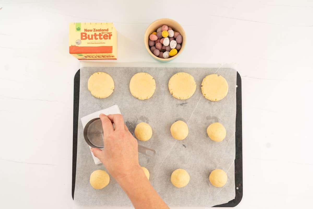 Balls of cookie dough on a baking sheet being flattened by a measuring cup in to round disk shaped cookies.