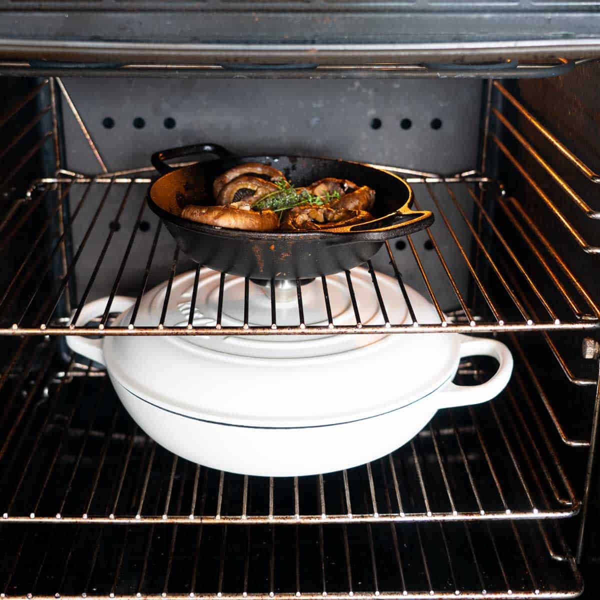 An oven containing a roasting dish of mushrooms and a large white enamel lidded dish.