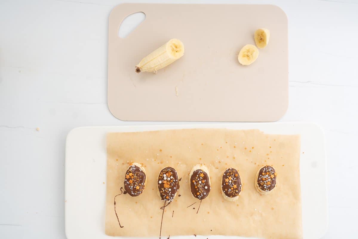 Chocolate covered banana slices decorated with golden sprinkles on a baking paper lined tray.