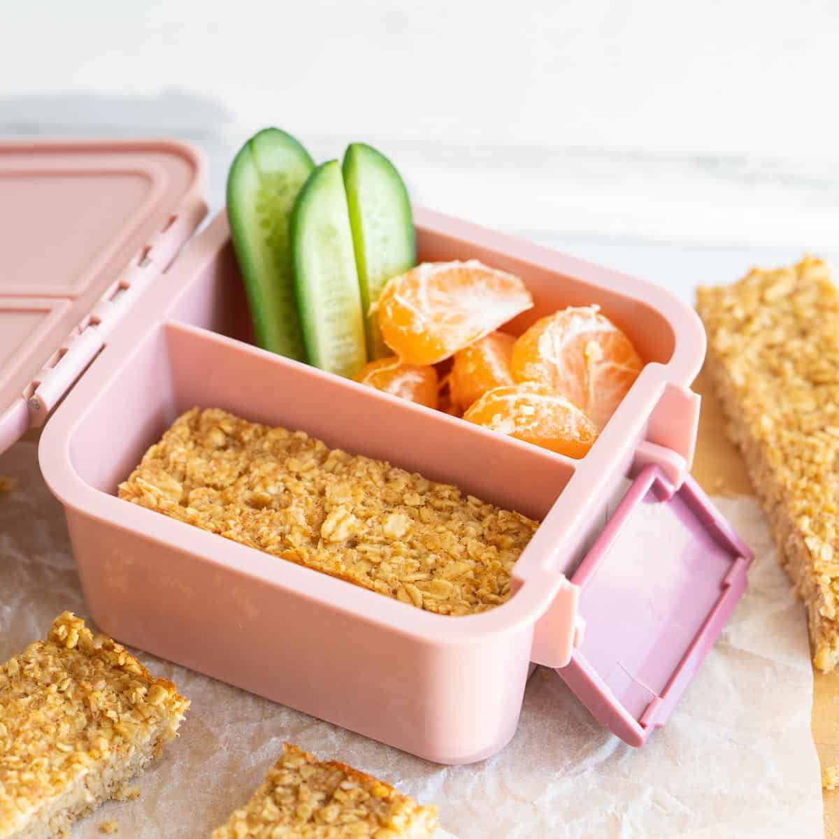 Oat bars packed into a pink bento lunch box with cucumber sticks and mandarin segments. 