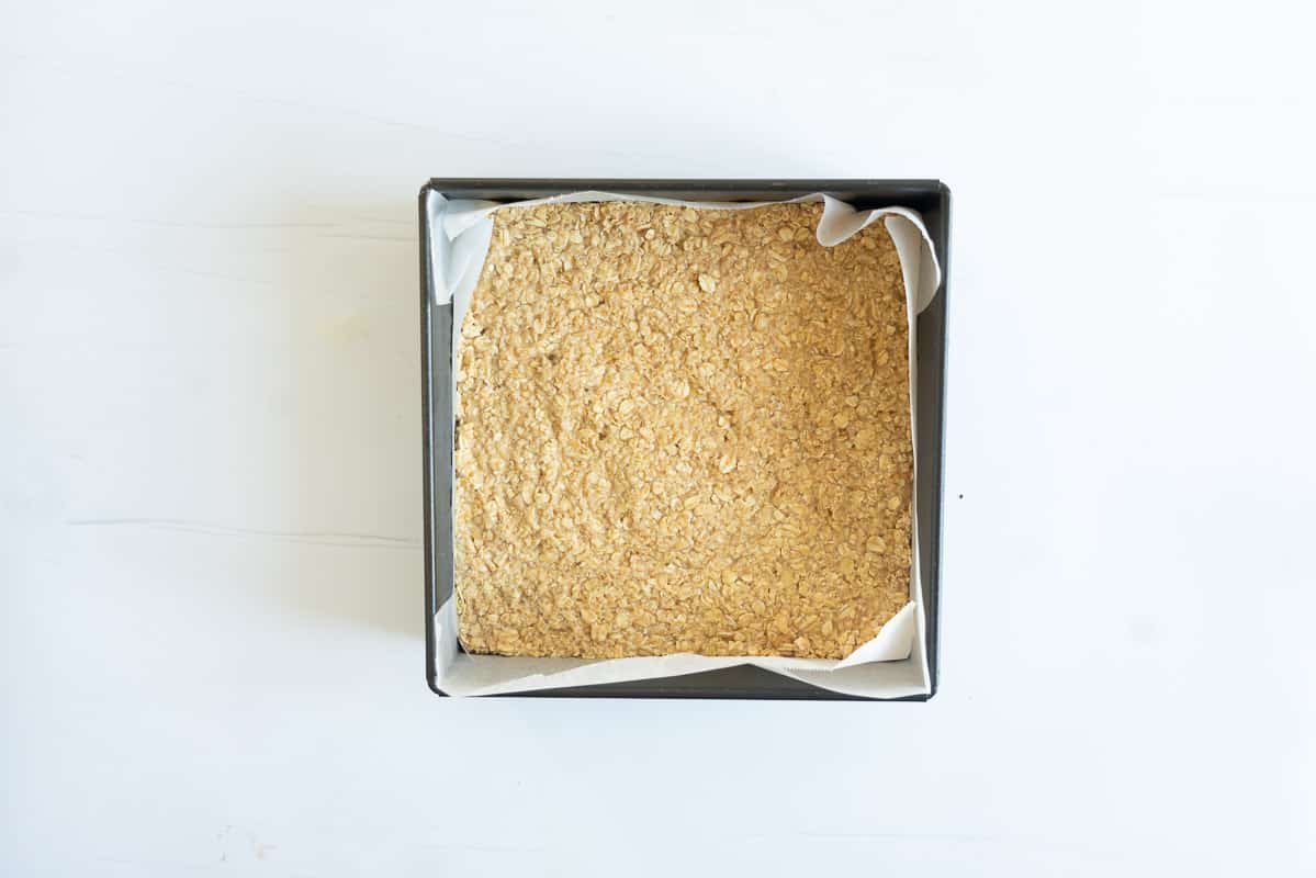 Oatmeal bar mix pressed into the bottom of a square lined cake tin.
