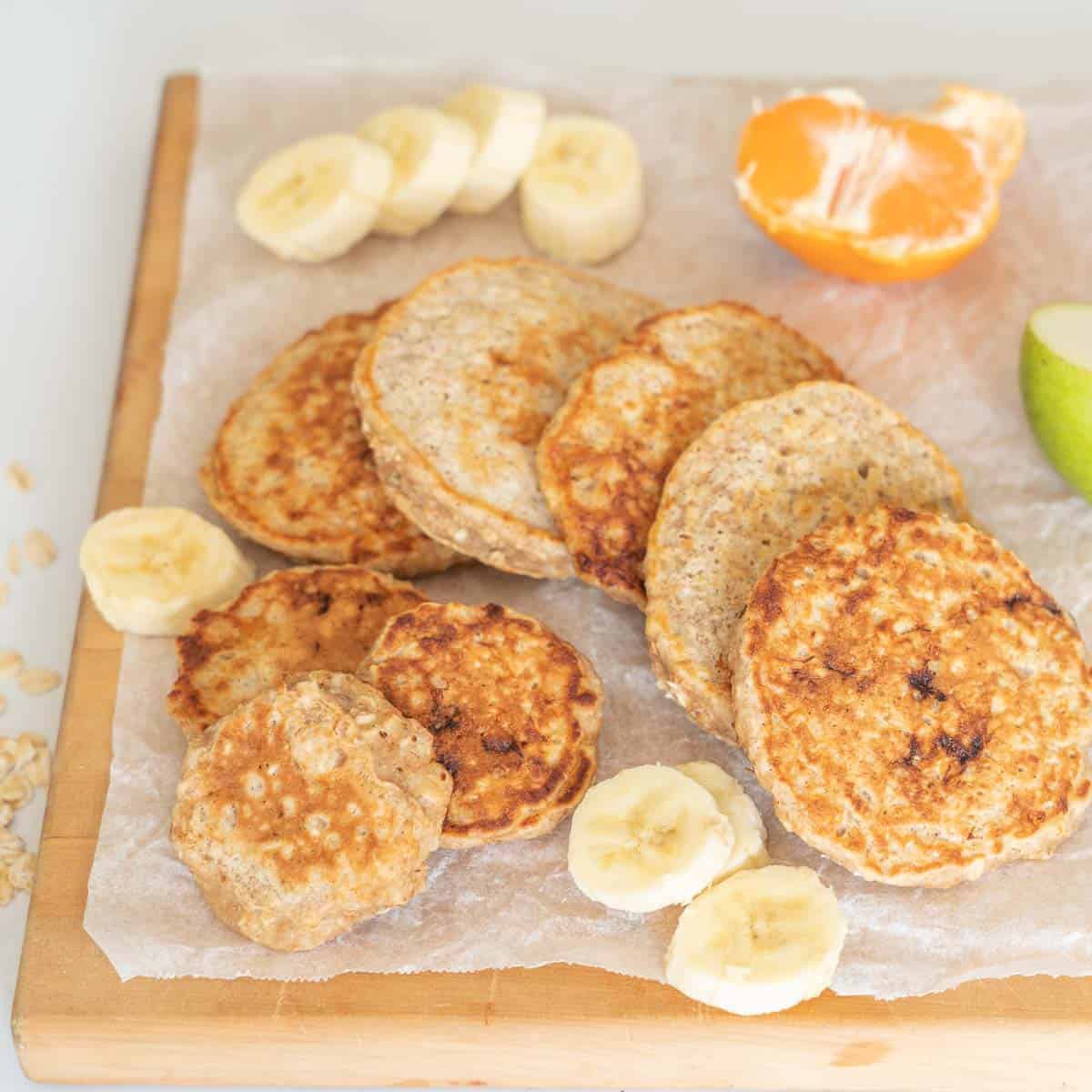 large and small banana pikelets on a wooden chopping board with cut fruit.