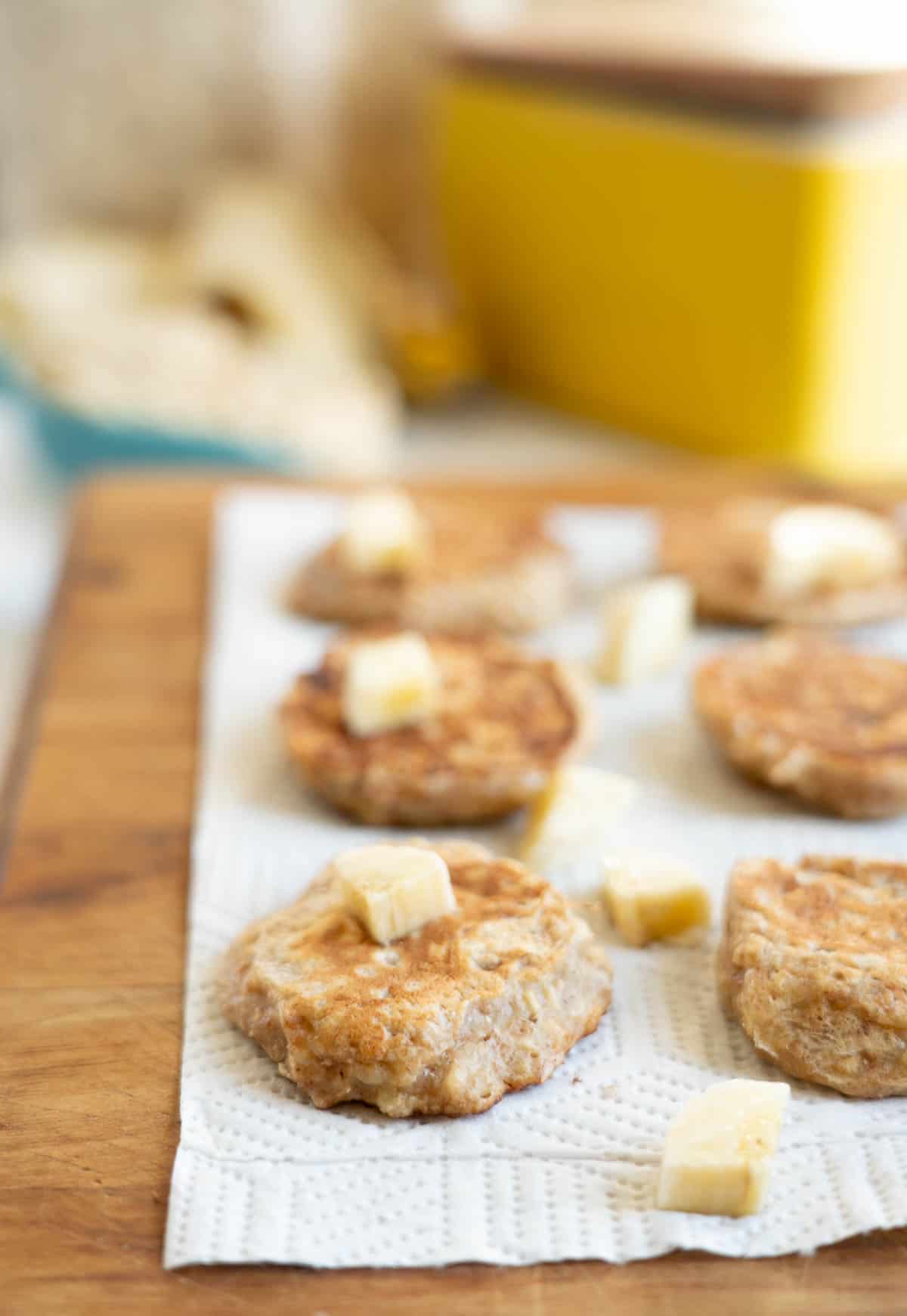 Banana oat pikelets on a paper towel scattered with small pieces of banana, a yellow butter dish in the background.