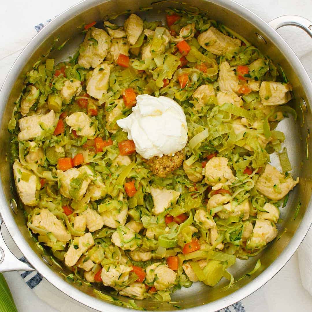 A spoonful of sour cream and whole grain mustard being added to cooked chicken and veggies.
