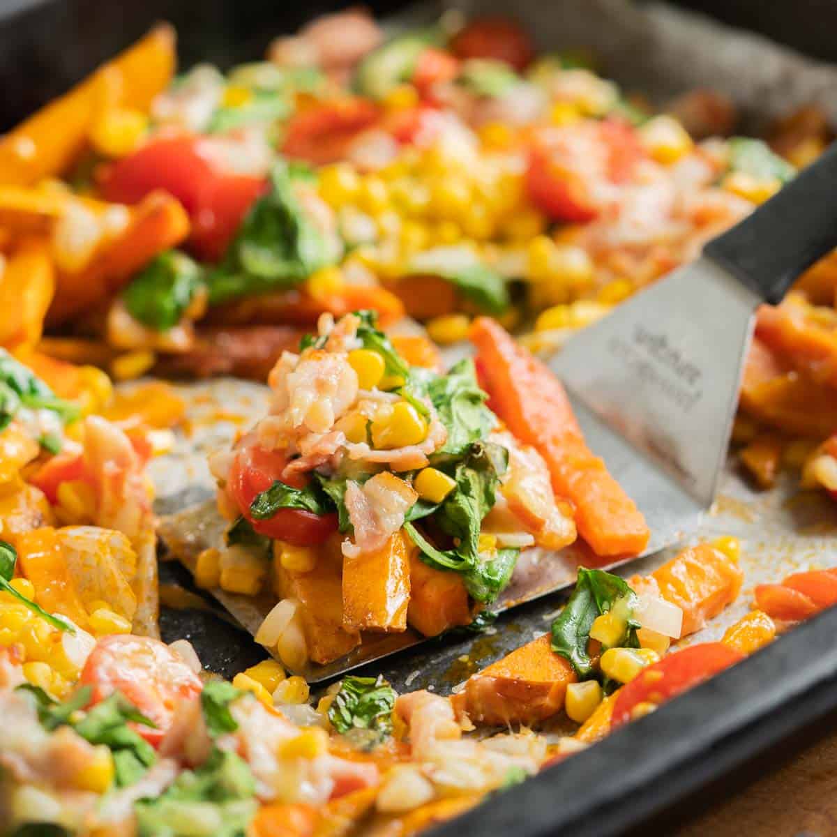 A large spatula lifting kumara battens topped with cooked vegetables, bacon and melted cheese from a large roasting pan.