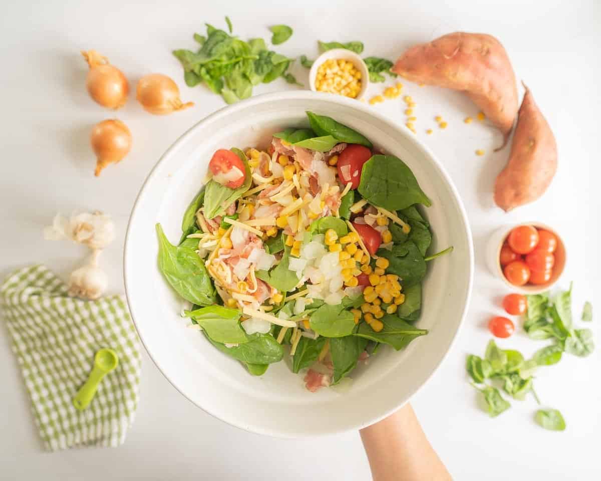 Colourful vegetables, grated cheese and chopped bacon in a white ceramic bowl.