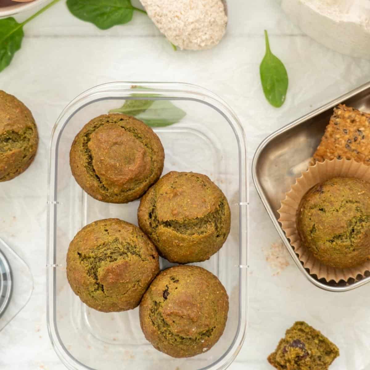 Four muffins in an airtight container next to a stainless steel lunch box packed with a muffin, crackers and fruit.