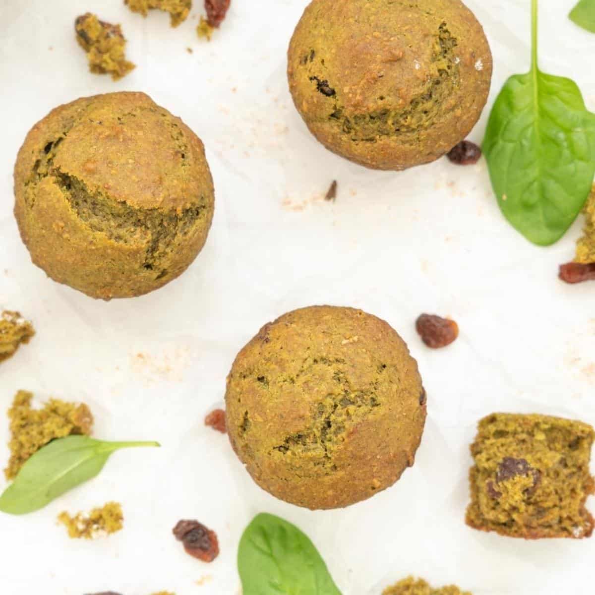 Green spinach muffins on crinkled baking paper scattered eith raisins and spinach leaves.
