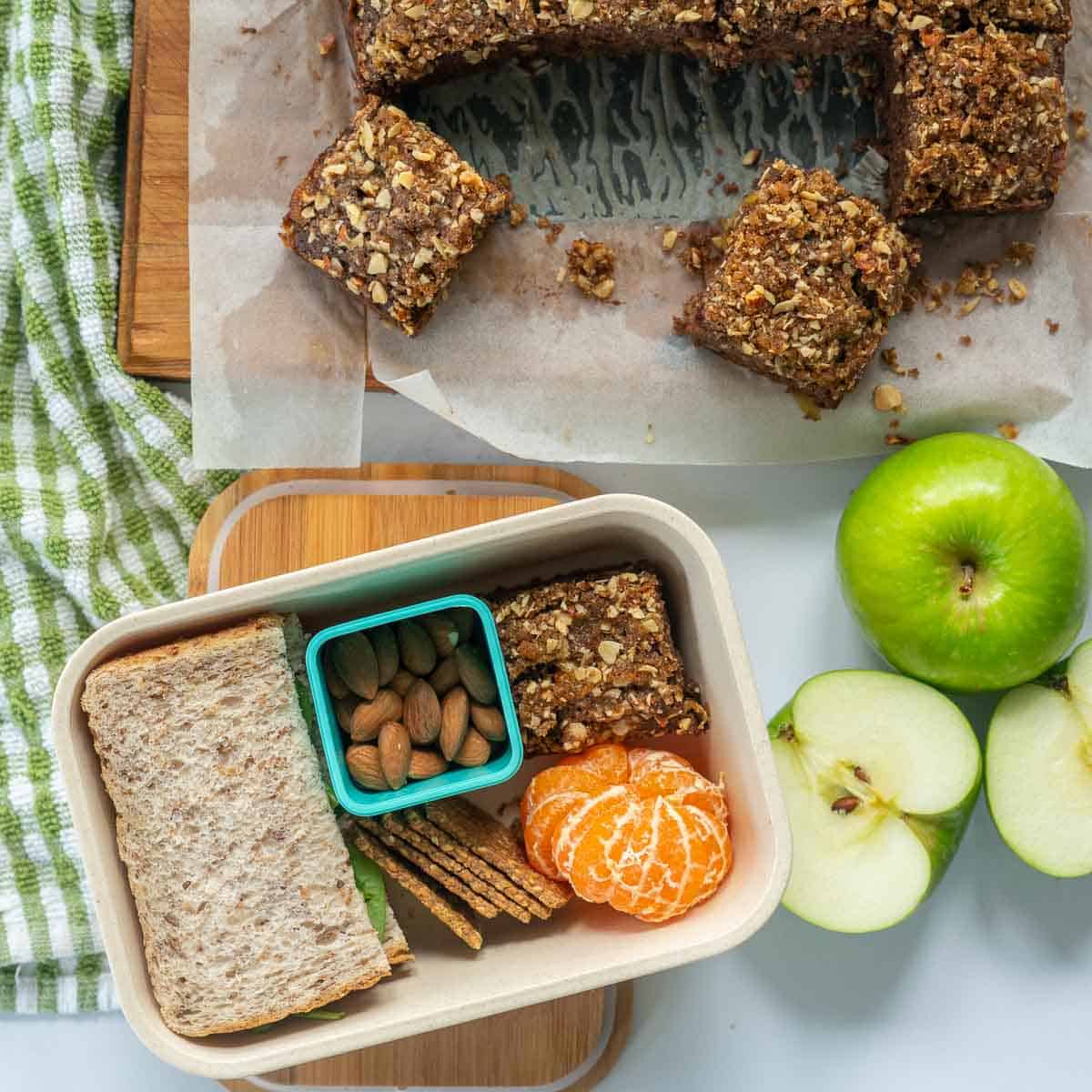 A lunchbox packed with a piece of apple cake, fruit, nuts, crackers and a sandwich.