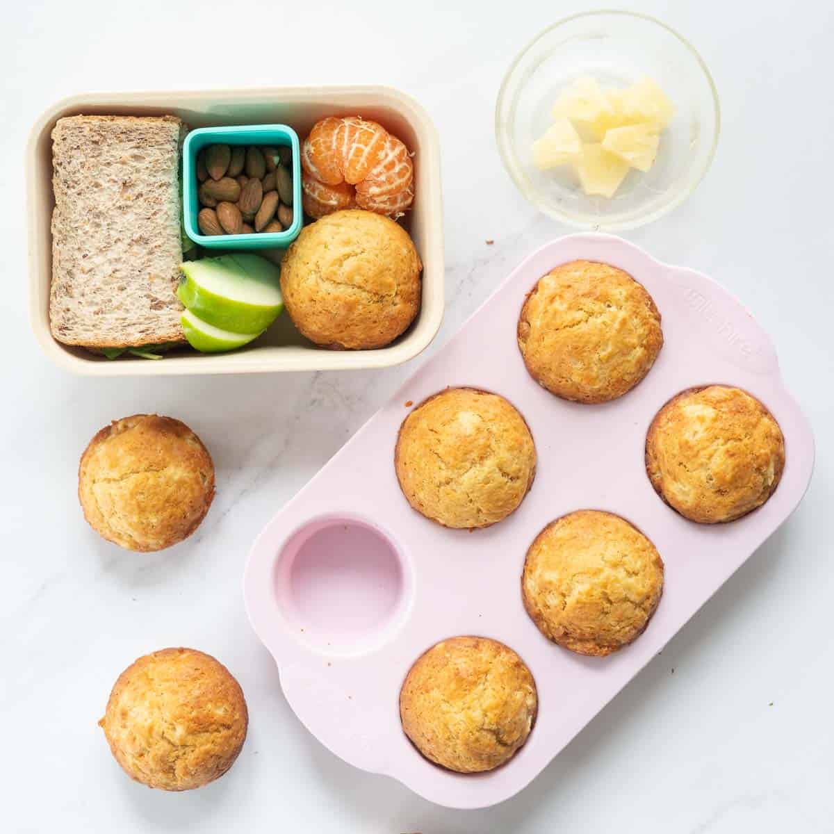 A pink muffin tray of muffins next to a packed shcool lunch box and a bowl of pineapple pieces. 