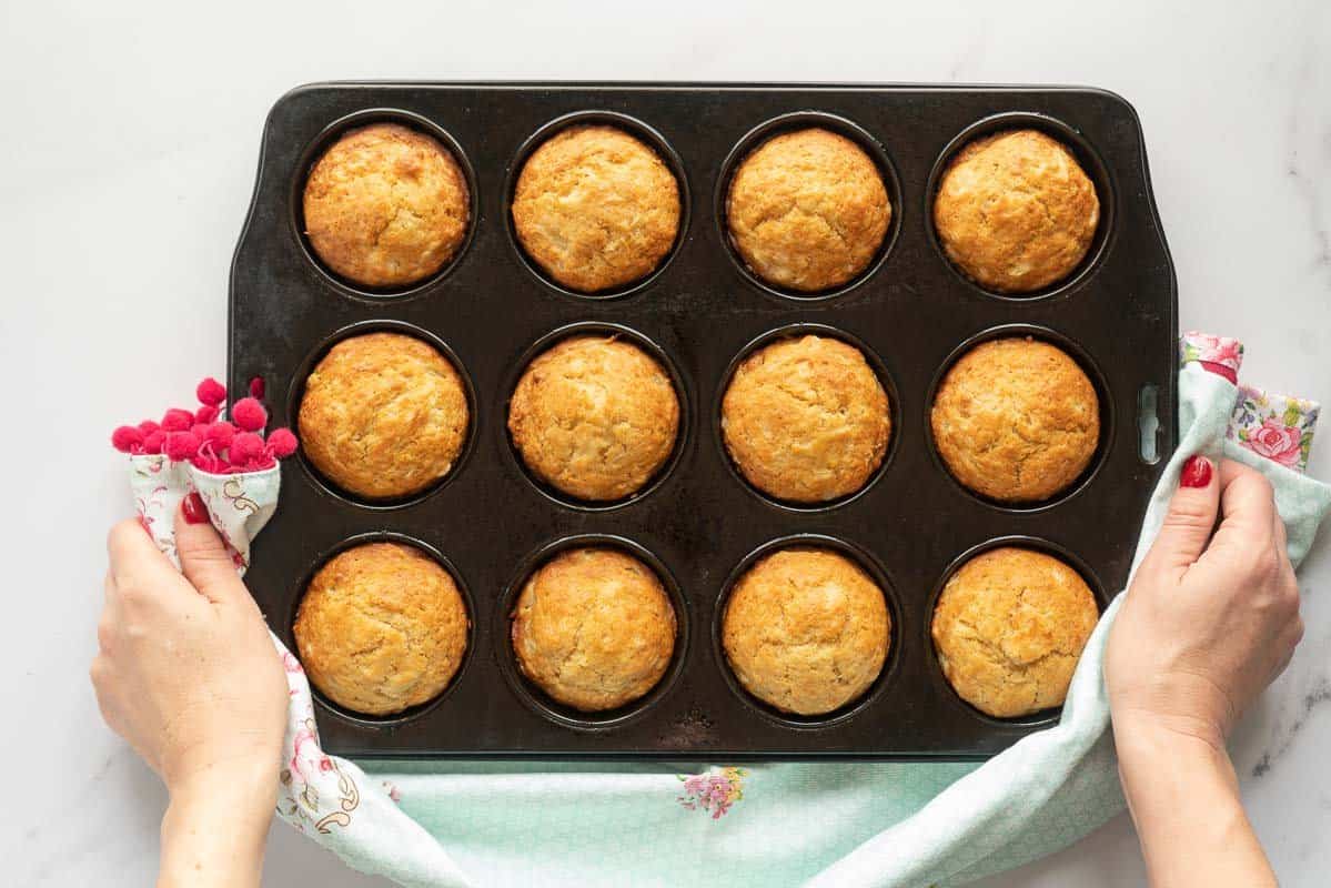 Golden baked muffins in a black muffin tray held with two hands using a floral tea towel. 