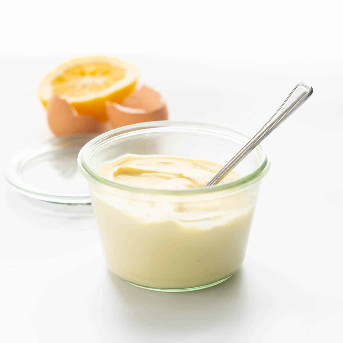 A glass jar filled with cream coloured mayonnaise, egg shells and half a lemon in the background.