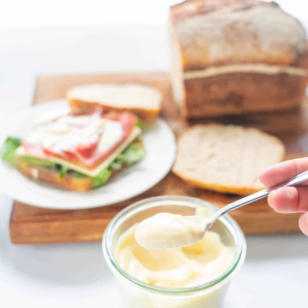 A spoonful of mayonnaise in front of a wooden board with a sandwich and loaf of bread.