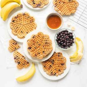 Three plates of waffles on a bench with a bowl of blueberries, maple syrup and bananas.