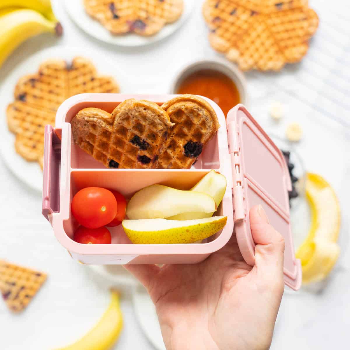 A small pink lunchbox picked with waffles, tomatoes and pears. 