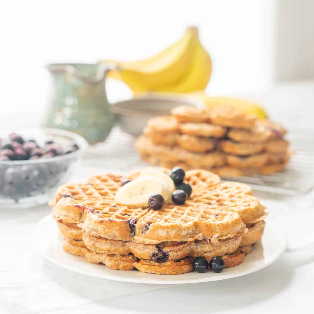 A stack of three waffles topped with banana slices and blueberries, a jug if maple syrup in the background.