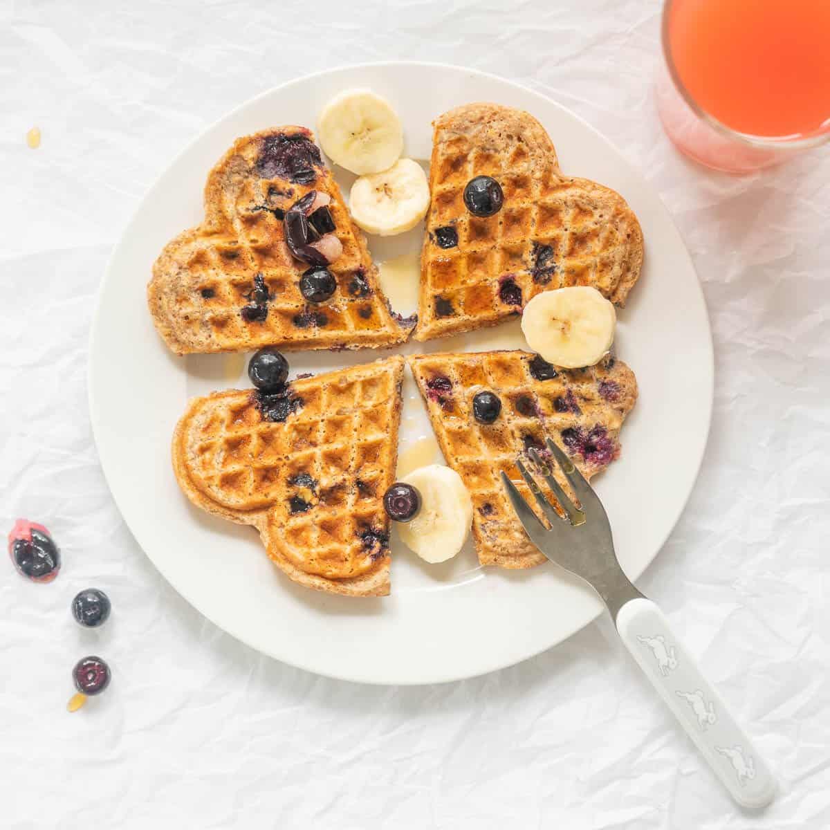 4 heart shaped waffles on a white plate scattered with banana slices and blieberries. 