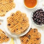 Three plates of waffles on a bench with a bowl of blueberries, maple syrup and bananas.